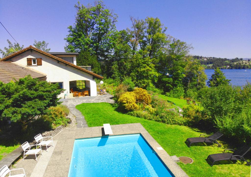 Experience the serene blend of luxury and nature at Classic Lakeside Villa Lucerne, nestled against the stunning backdrop of the Swiss Alps. This image captures the villa's majestic charm, its inviting infinity pool, and the pristine waters of Lake Lucerne, offering guests a picturesque retreat.